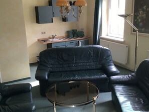 A living room with a black leather sofa, a glass coffee table, and a kitchenette area with a stove and cabinets.
