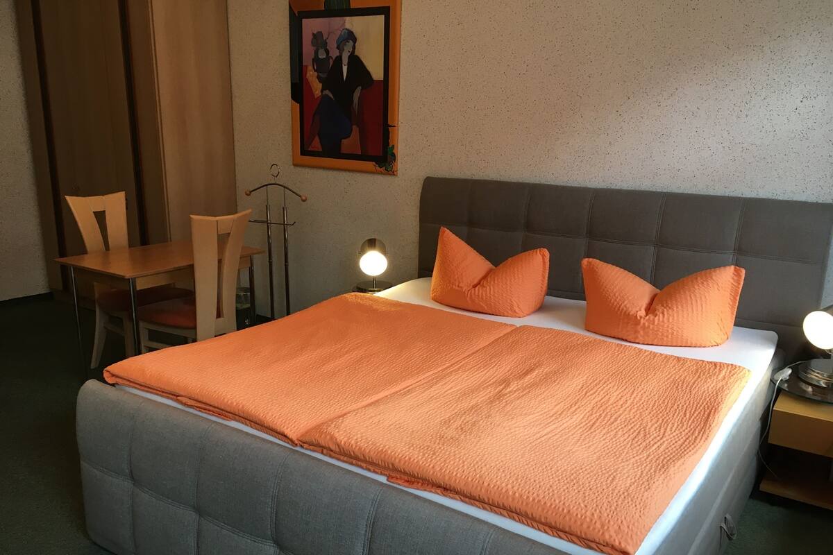A neatly made bed with an orange blanket, a grey upholstered headboard, and two bedside lamps.