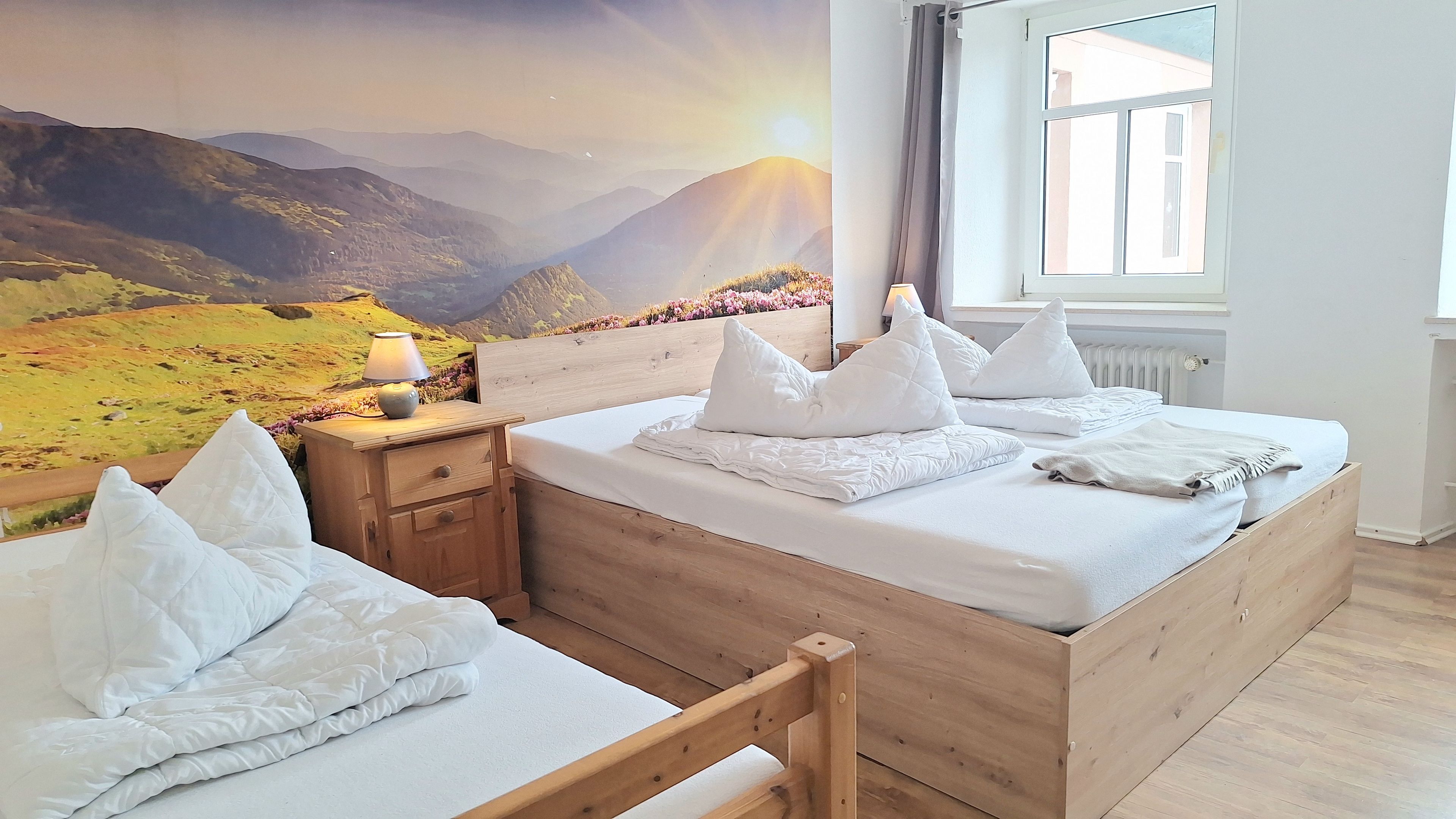 Une chambre avec un lit en bois, du linge de lit blanc, une table de chevet, une lampe et une fresque murale représentant un paysage de montagne.