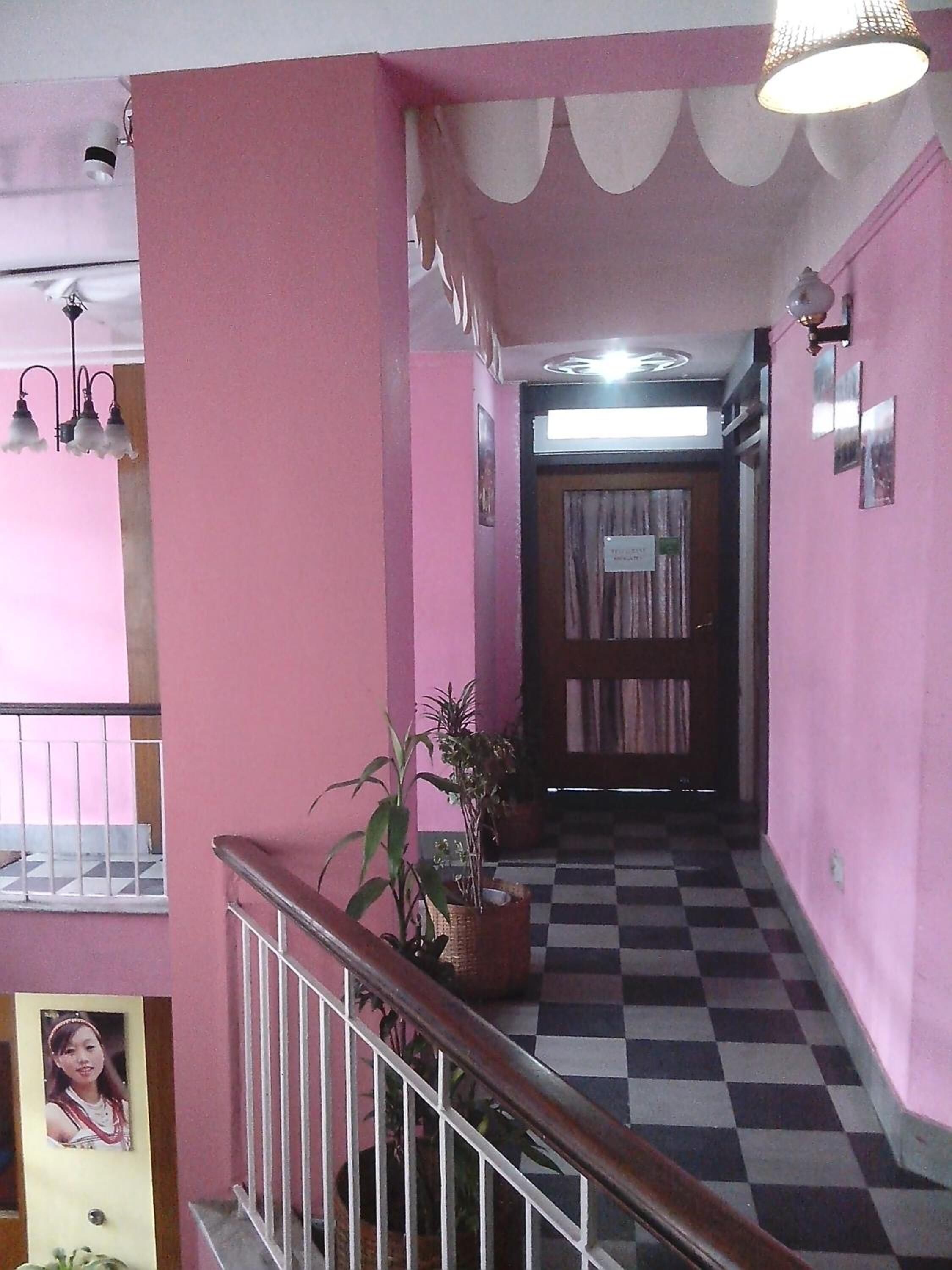 Un pasillo de paredes rosadas con piso a cuadros, una escalera con barandilla y una planta en maceta.
