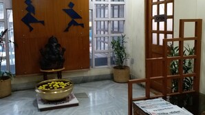 Una habitación con puerta de madera, un ventanal grande, una estatua de Buda, un cuenco de flores y una silla de madera con un periódico.