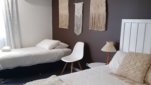 A hotel room with a large bed, two smaller beds, a white chair, macrame wall hangings, an air conditioning unit, and a lamp. - Town House Motor Inn (Young)