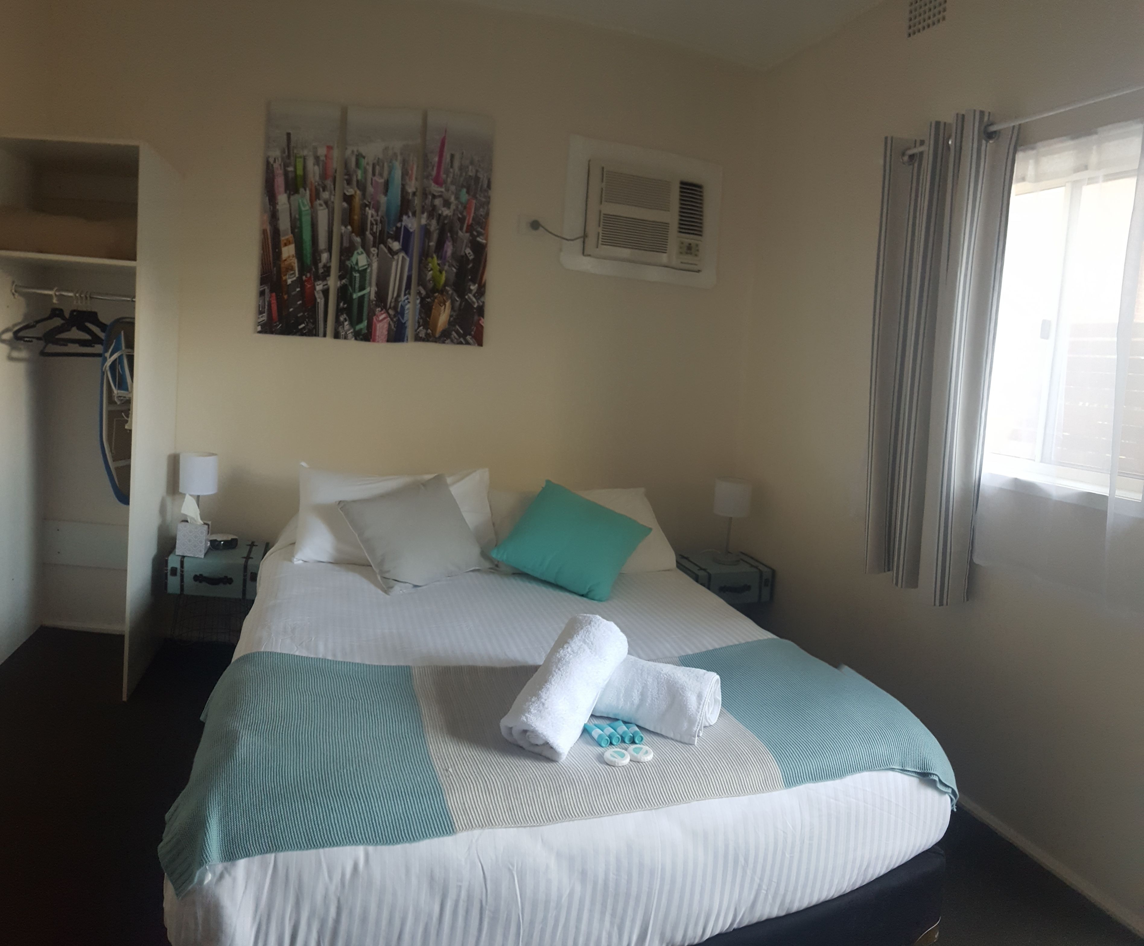 A hotel room with a bed, a painting on the wall, an air conditioning unit, a window with curtains, and a coat rack with clothes.