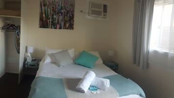 A hotel room with a bed, a painting on the wall, an air conditioning unit, a window with curtains, and a coat rack with clothes.