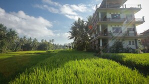 A multi-story building with balconies, surrounded by lush green fields and palm trees. - Nirwa Ubud Karma (Ubud)