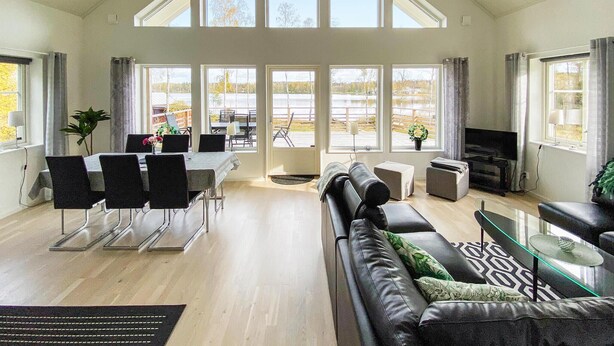 A spacious living area with a high ceiling, large windows, a dining area with chairs and a table, a sofa, a TV, and a small table with a lamp.