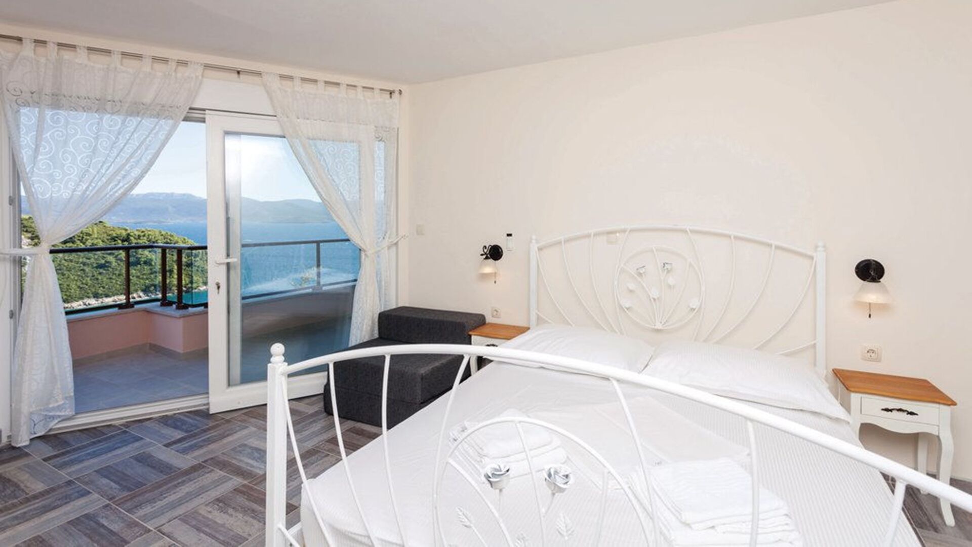 Un dormitorio con una cama blanca, una mesita de noche pequeña, un buró y vistas a un balcón con barandilla, además de una puerta que da a un espacio exterior.
