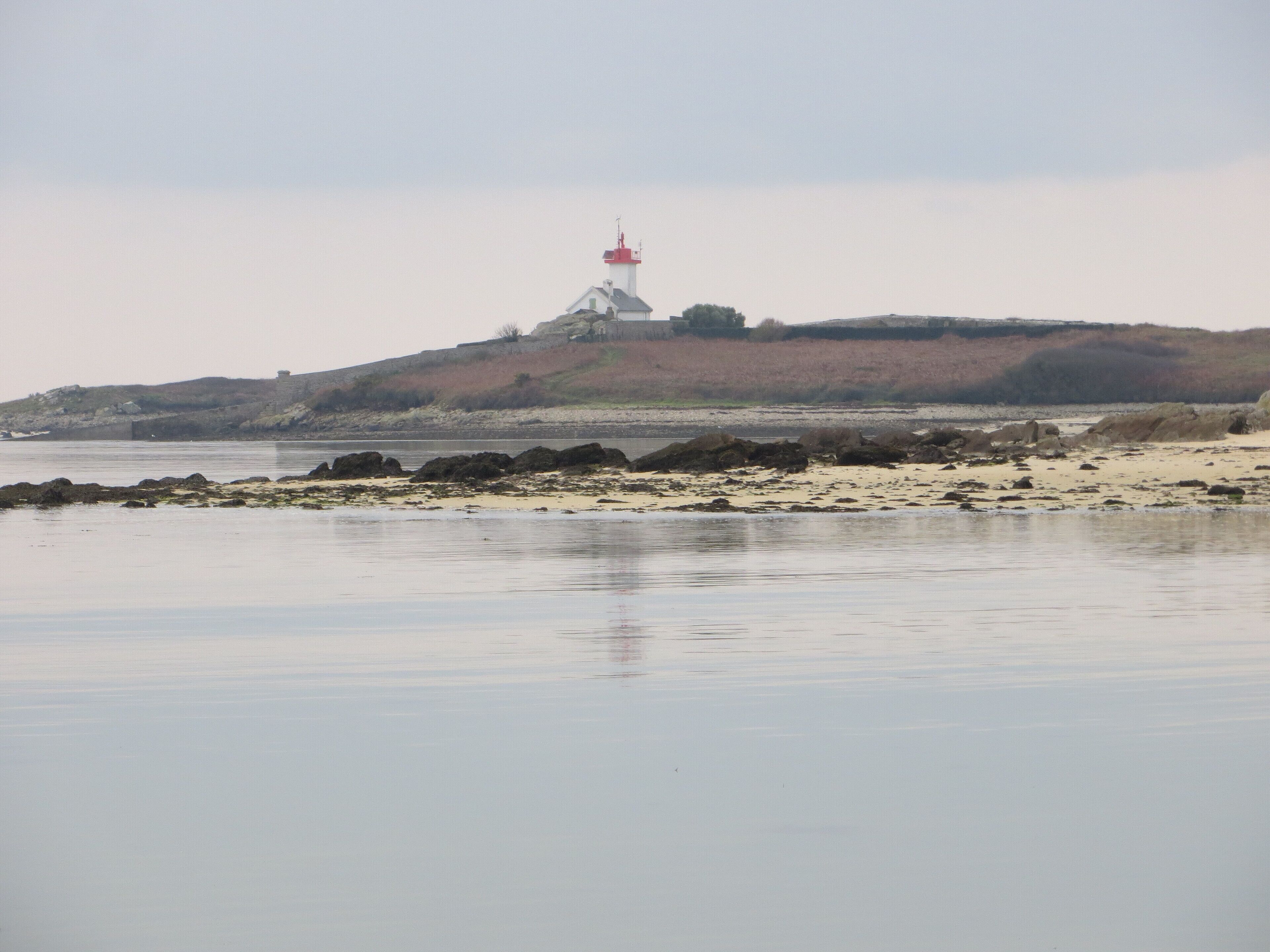 Paisagem costeira com um farol no topo de uma colina, águas calmas e costa rochosa.