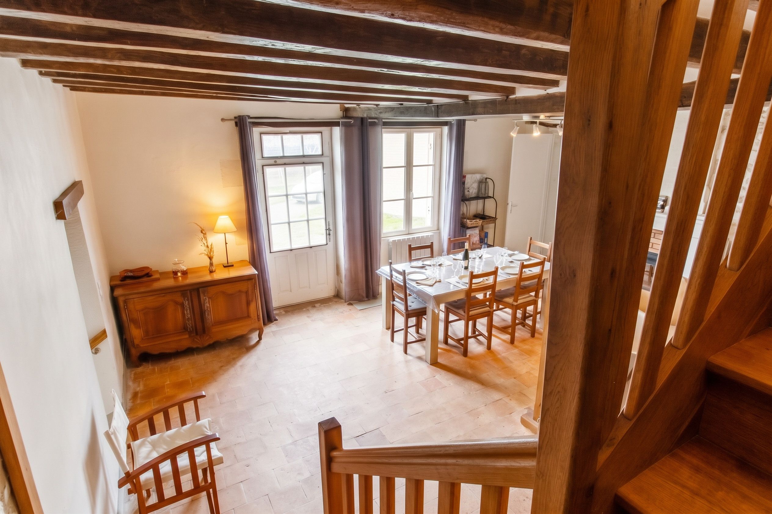 Un coin repas avec une table en bois dressée pour quatre personnes, un buffet en bois et une fenêtre avec des rideaux.