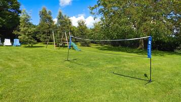 Ein grasbewachsenes Freigelände mit Volleyballnetz, Schaukeln und einer blauen Flagge.