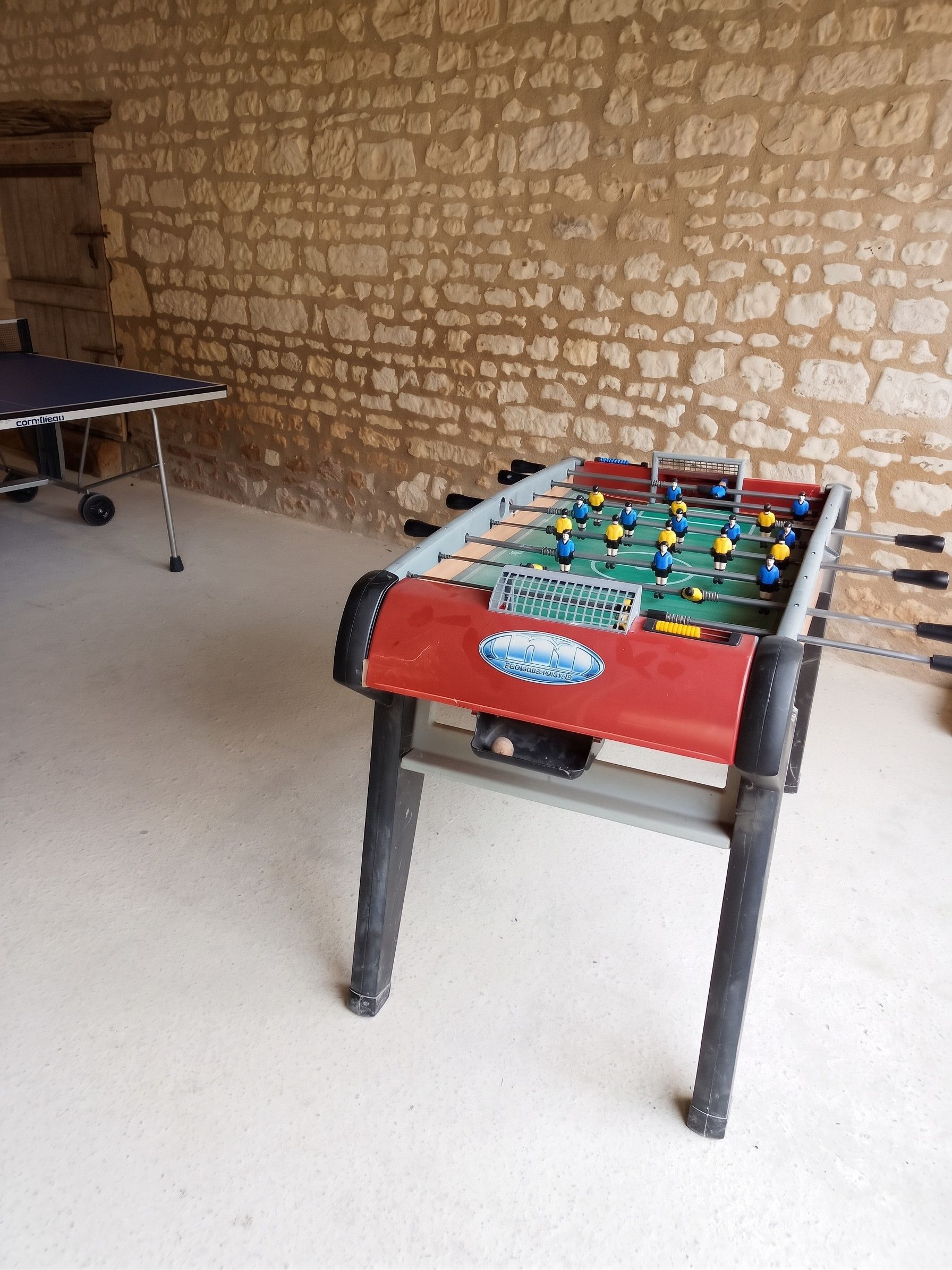 A foosball table in a room with a stone wall and a ping pong table in the background.