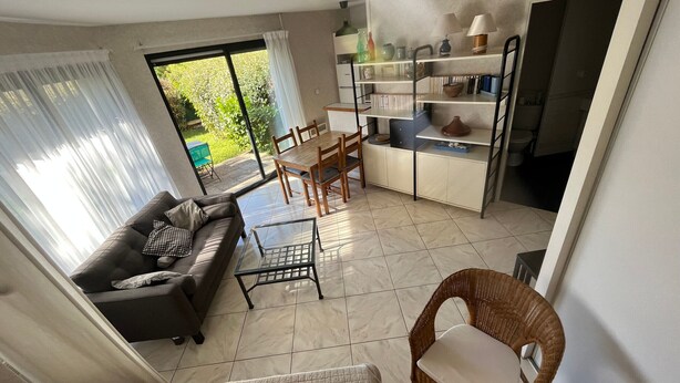 Una sala de estar con un sofá, una mesa de centro de vidrio, una mesa de comedor con sillas, una zona de cocina con estanterías y vistas a zonas verdes.