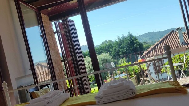 Quarto com cama, janela com vista para uma casa e vegetação, e varanda com guarda-corpo.