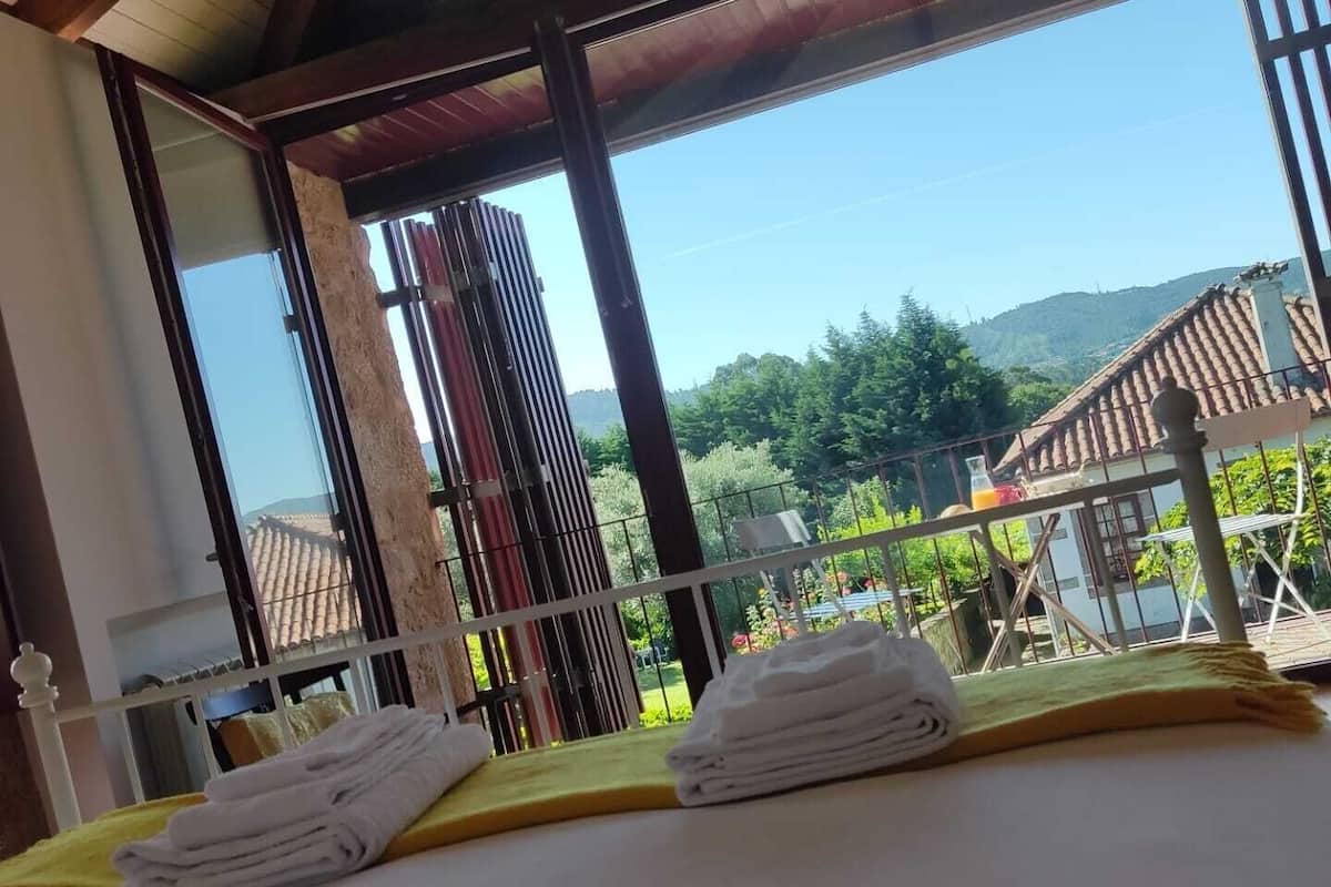 A room with a bed, a window with a view of greenery and buildings, and a balcony with a railing.