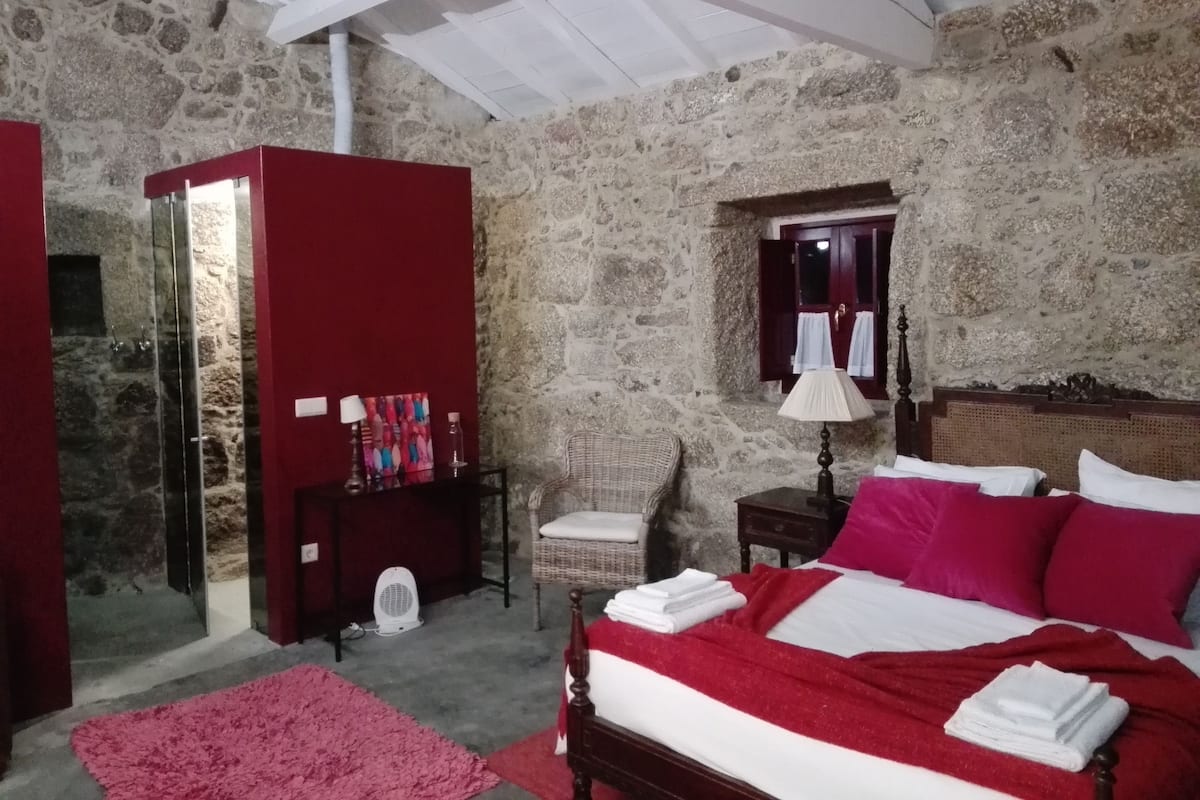 A bedroom with stone walls, a bed with white and red bedding, a wooden nightstand, a wicker chair, and a red wardrobe.
