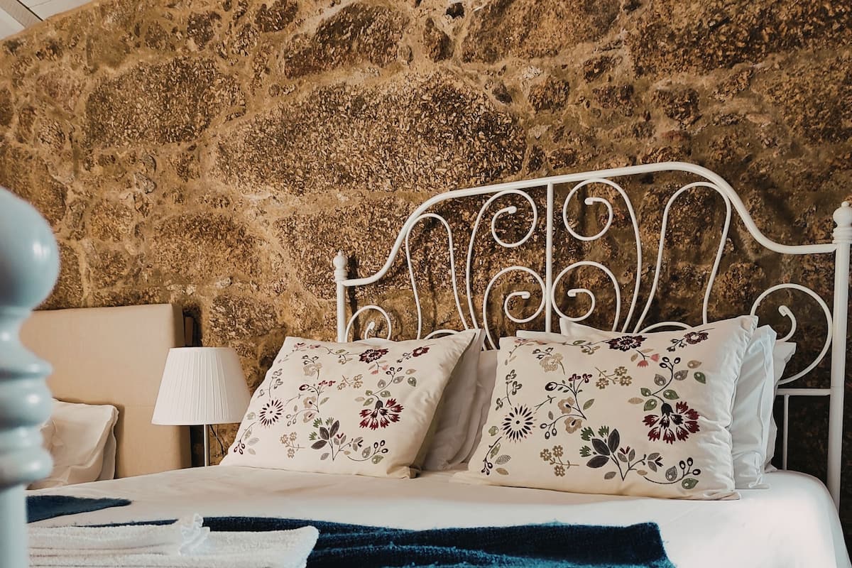 A bed with white bedding and floral pillows, a blue blanket, and a stone wall behind.