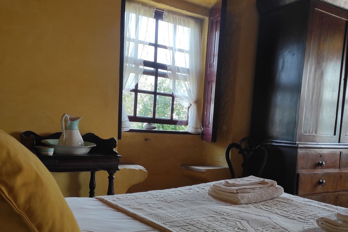 A hotel room with a bed, a desk with a pitcher, a window with curtains, and a wardrobe.