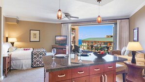 A hotel room with a bed, a dining area with a table and chairs, a TV, and a view of a building and the sea. - Maui Resort Rentals: Honua Kai Konea 815 - Stunning 8th Floor Ocean View Studio (Lahaina)