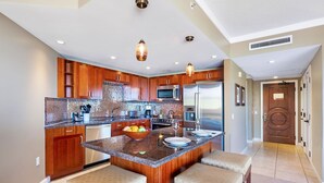 A modern kitchen with wooden cabinets, a central island with seating, stainless steel appliances, and a tiled backsplash. - Maui Resort Rentals: Honua Kai Konea 815 - Stunning 8th Floor Ocean View Studio (Lahaina)