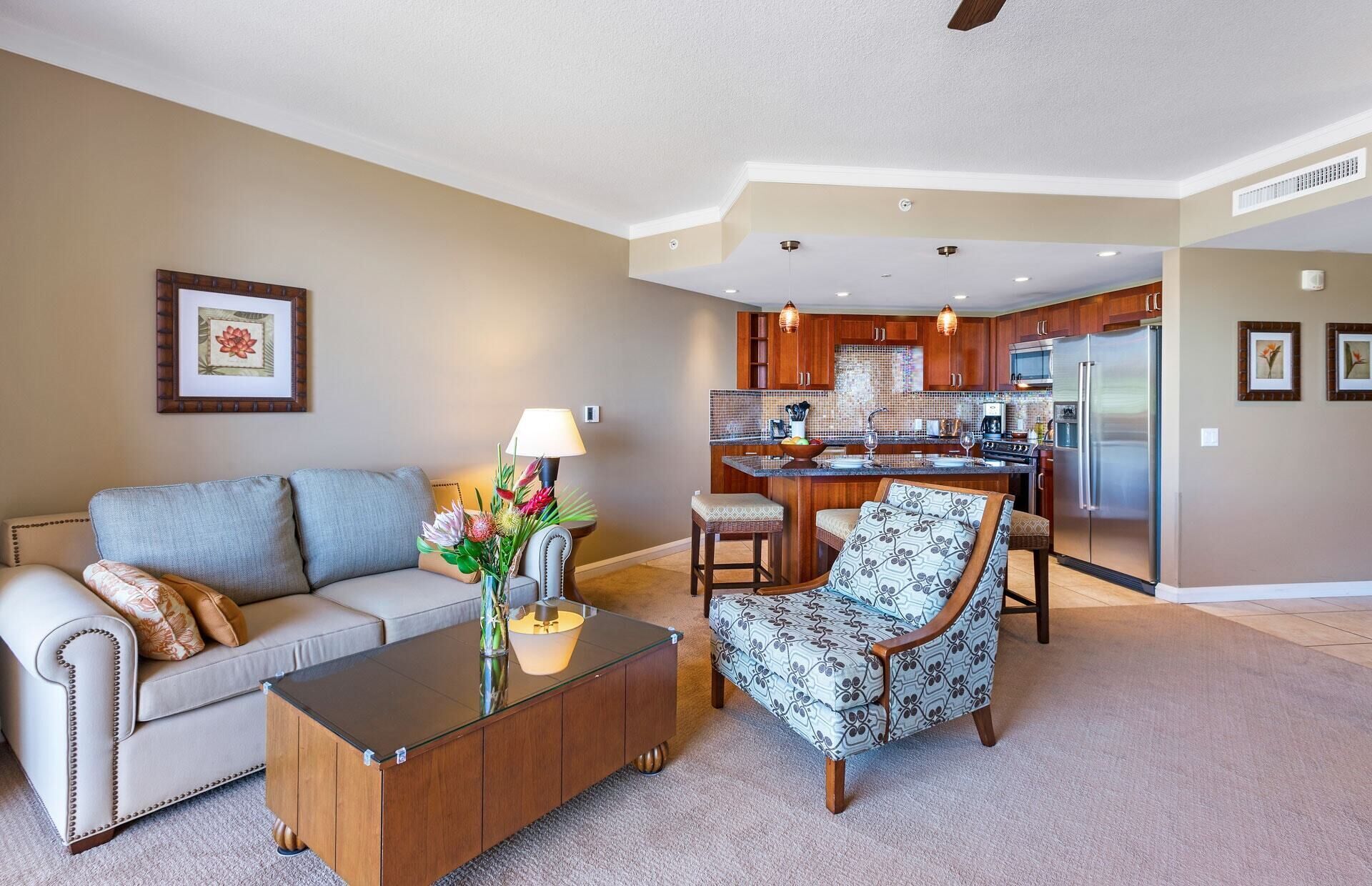 A living room with a sofa, a glass coffee table, and a patterned armchair. There is a kitchen area with wooden cabinets and a refrigerator.