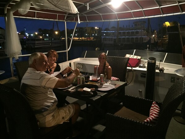 A dining area with a table and chairs, a person holding a drink, and a view of the water and boats in the background.