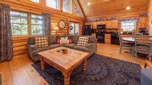 A log cabin interior with a living room, sofa set, wooden coffee table, kitchen area, and large windows. - Deep Creek Gem w/ Indoor Hot Tub (OAKLAND)