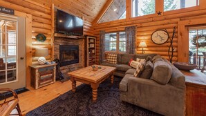A cozy log cabin interior with a fireplace, a television, a sofa, and a dining area. - Deep Creek Gem w/ Indoor Hot Tub (OAKLAND)
