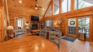 A log cabin interior with a wooden dining table, chairs, a fireplace, a television, and large windows. - Deep Creek Gem w/ Indoor Hot Tub (OAKLAND)