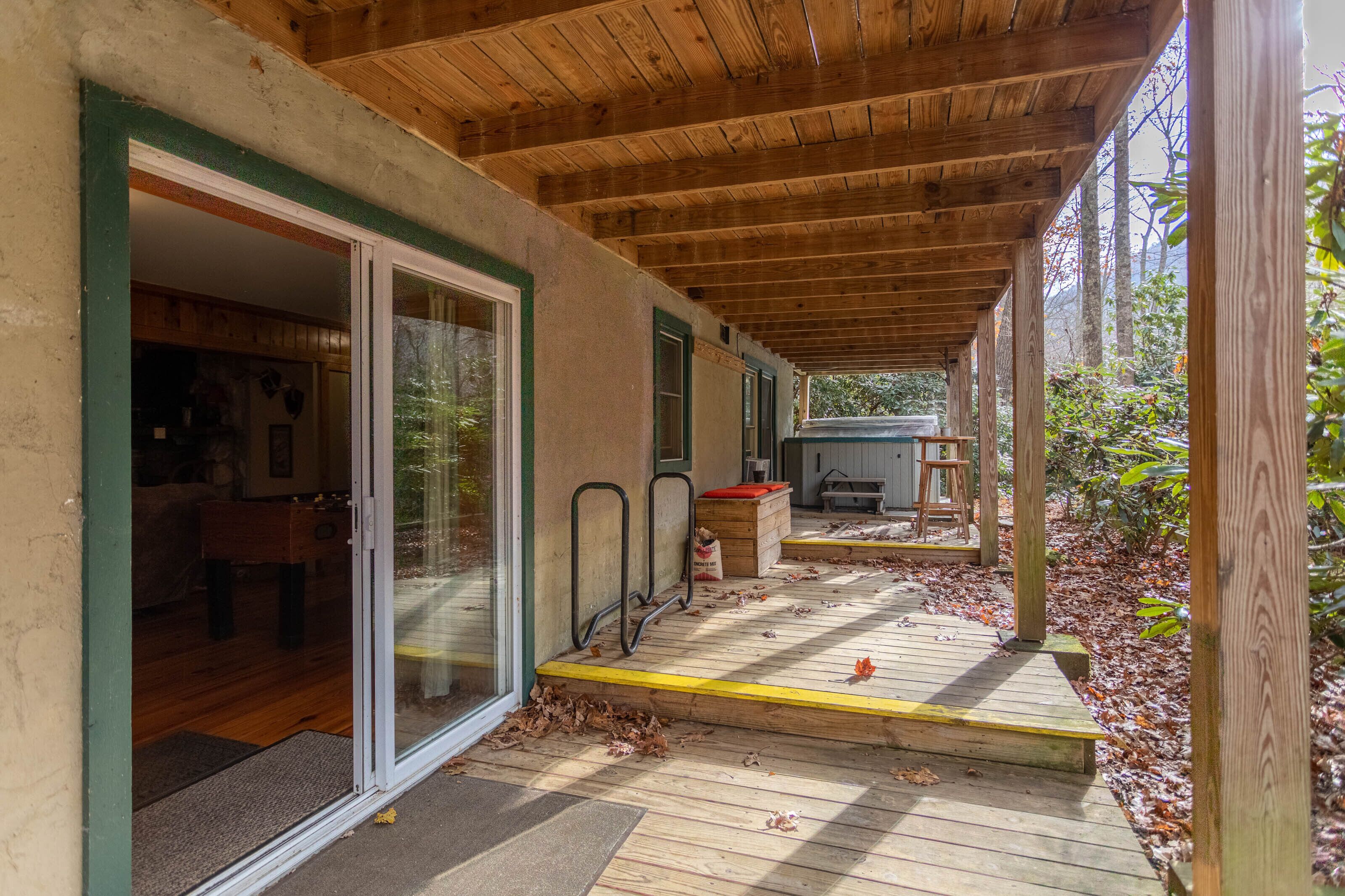 Une terrasse en bois couverte, avec une porte vitrée donnant sur une pièce, le tout entouré d’un jardin agrémenté de marches en bois et d’un banc.