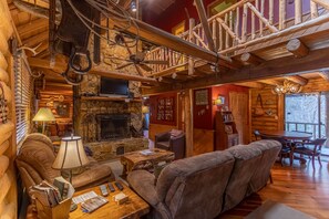 Interior de una cabaña de troncos con chimenea de piedra, mobiliario de cuero y una escalera de madera.