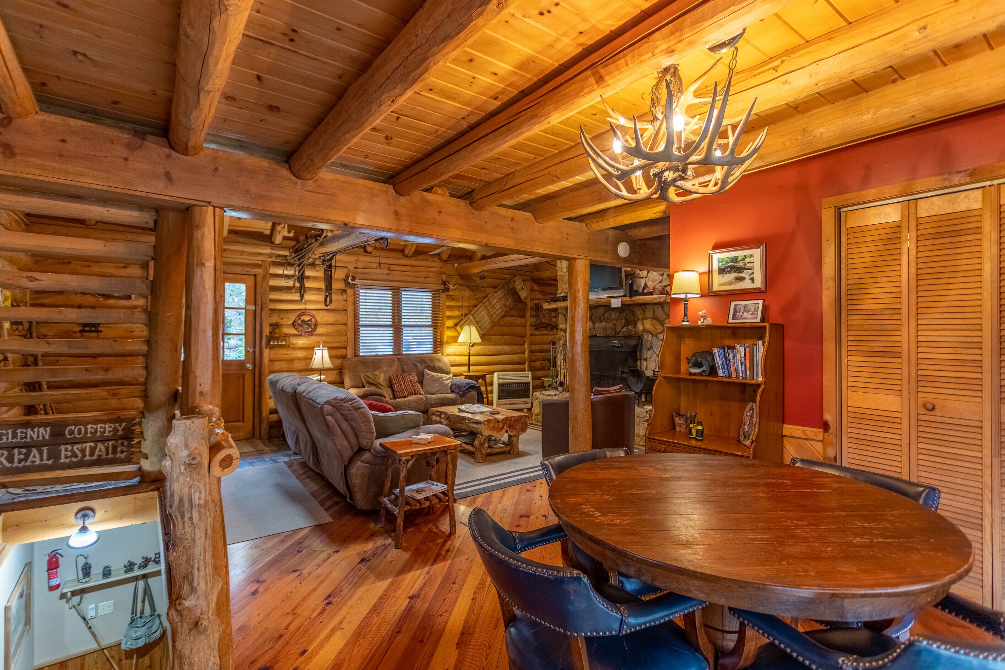 A log cabin interior with a wooden dining table, leather chairs, a fireplace, and a chandelier.