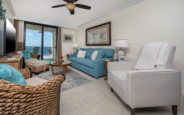 A living room with a blue sofa, a grey armchair, wicker furniture, and a view of the ocean through a sliding door.