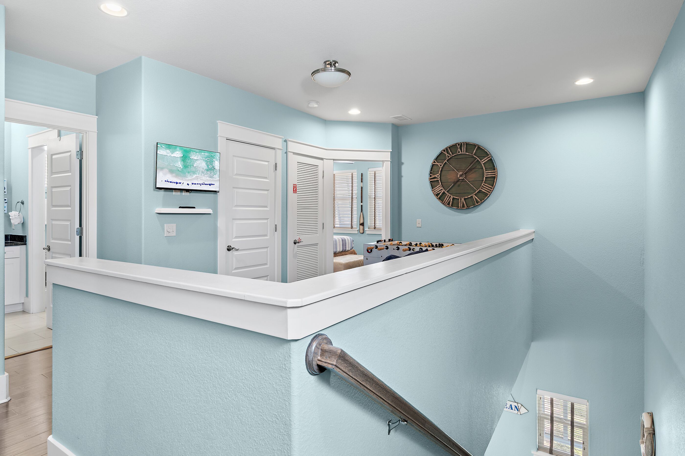 A modern interior with a light blue wall, a round wall clock, a flat-screen TV, and a staircase with a metal handrail.