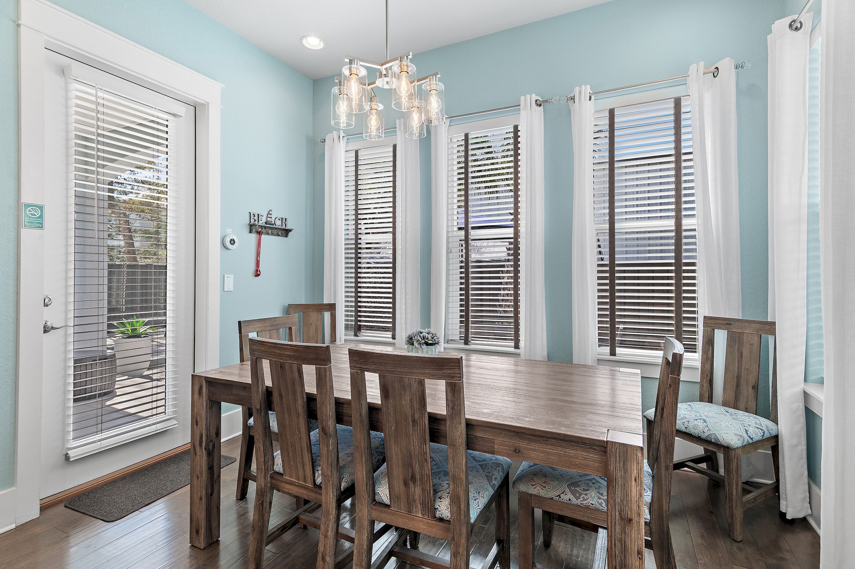 Comedor con mesa y sillas de madera, una lámpara de araña y ventanas con persianas.