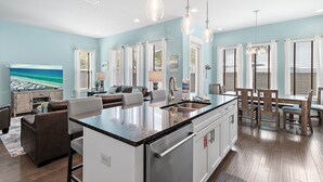 A modern kitchen with a central island, dining area with wooden chairs, and a living room with a sofa and TV. - Splash Pad⛳️Complimentary Golf Cart & New Pool 2026 | Sleeps 13 (Miramar Beach)