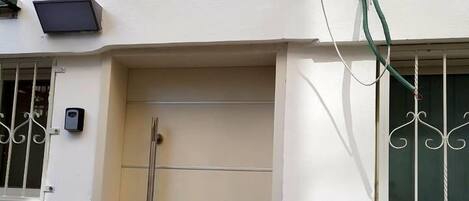 Una puerta blanca con manija de metal, un ventanal pequeño con celosías decorativas y una manguera verde a la derecha.