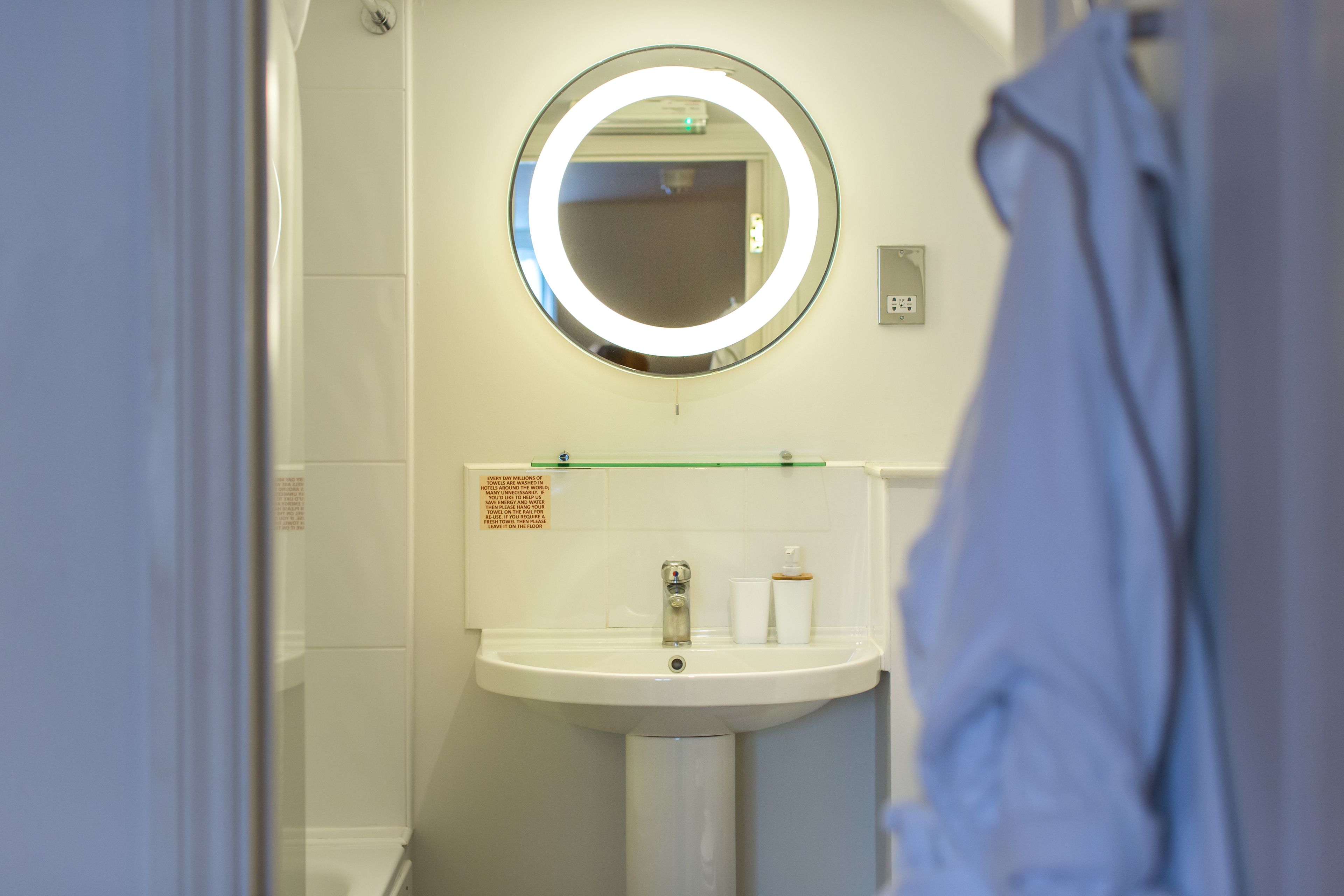 A bathroom with a round mirror, a sink, and a glass shelf with toiletries.