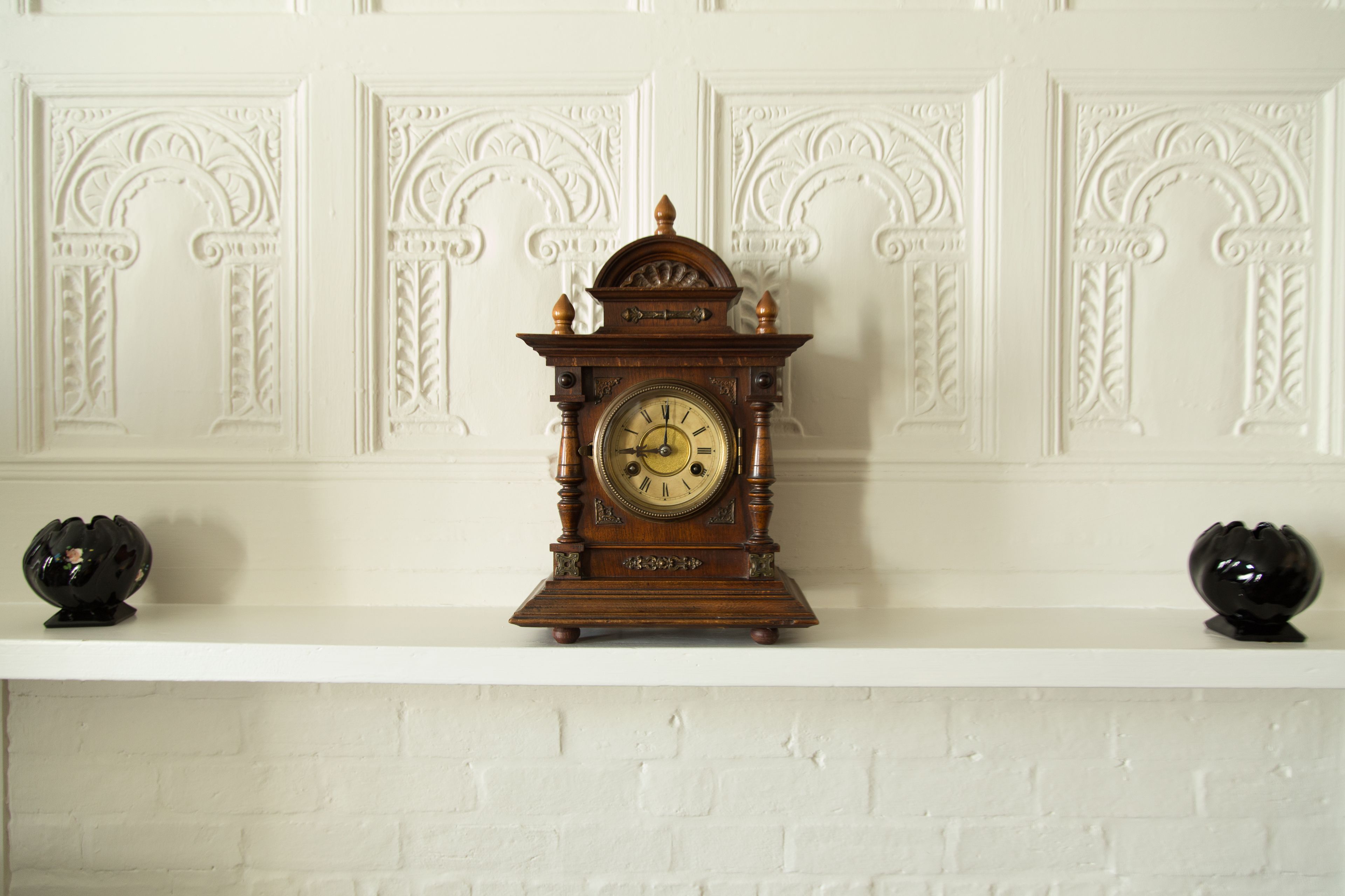 A wooden grandfather clock on a white mantelpiece against a white brick wall with decorative molding.