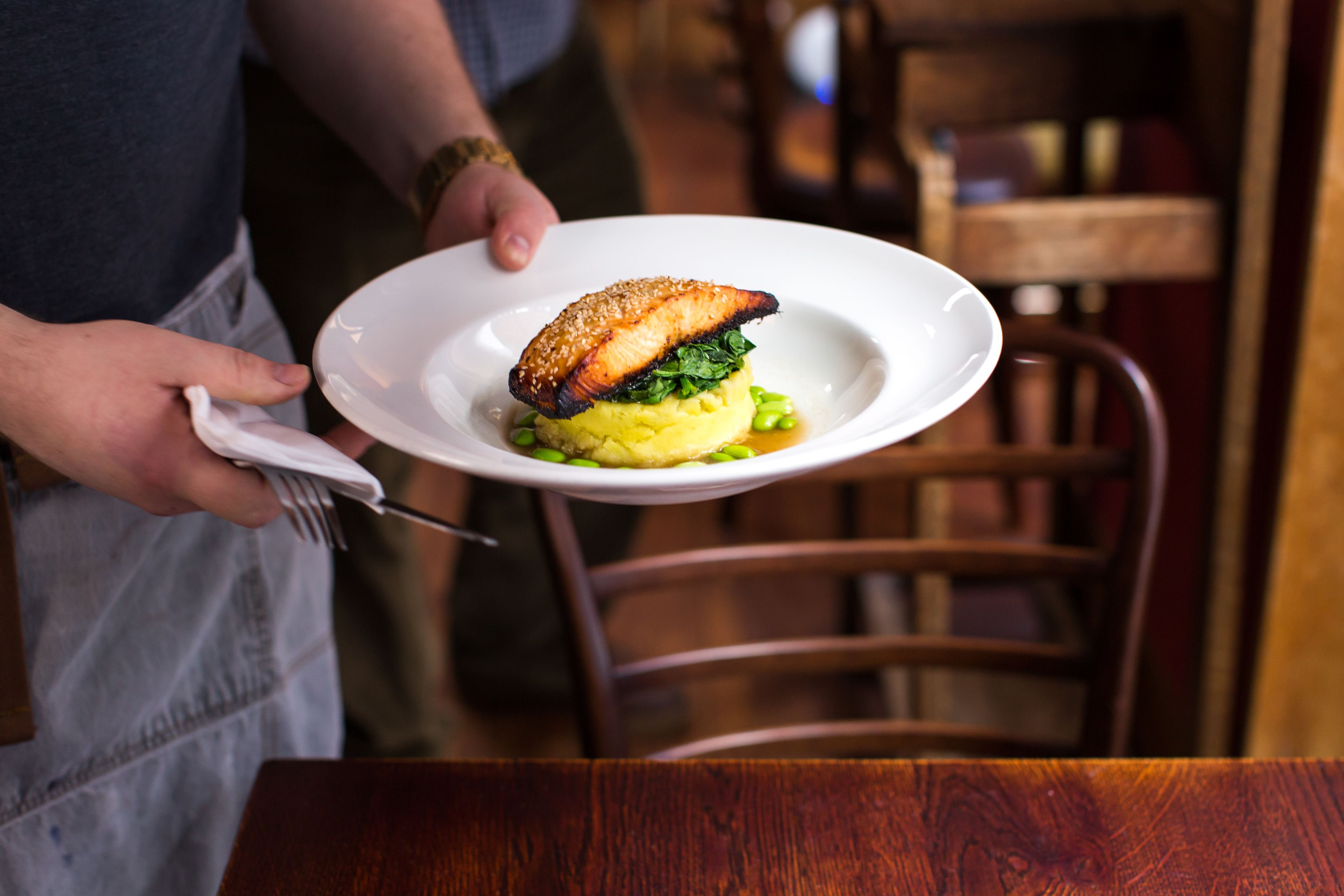 A person holding a white plate with a fish, greens, and yellow base, likely a dish, in a restaurant setting.