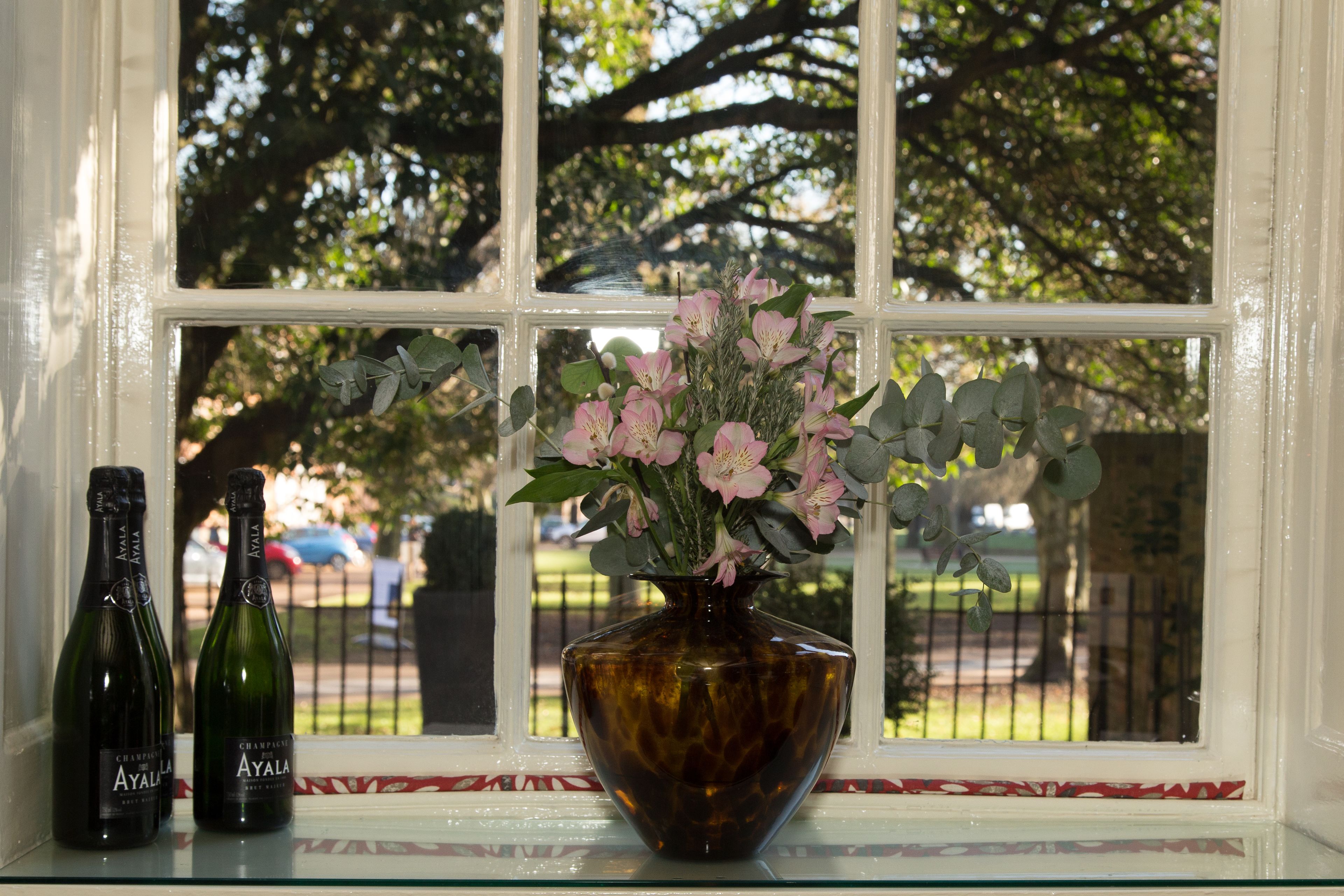 Une fenêtre avec un vase de fleurs et deux bouteilles de champagne Ayala sur une étagère en verre.