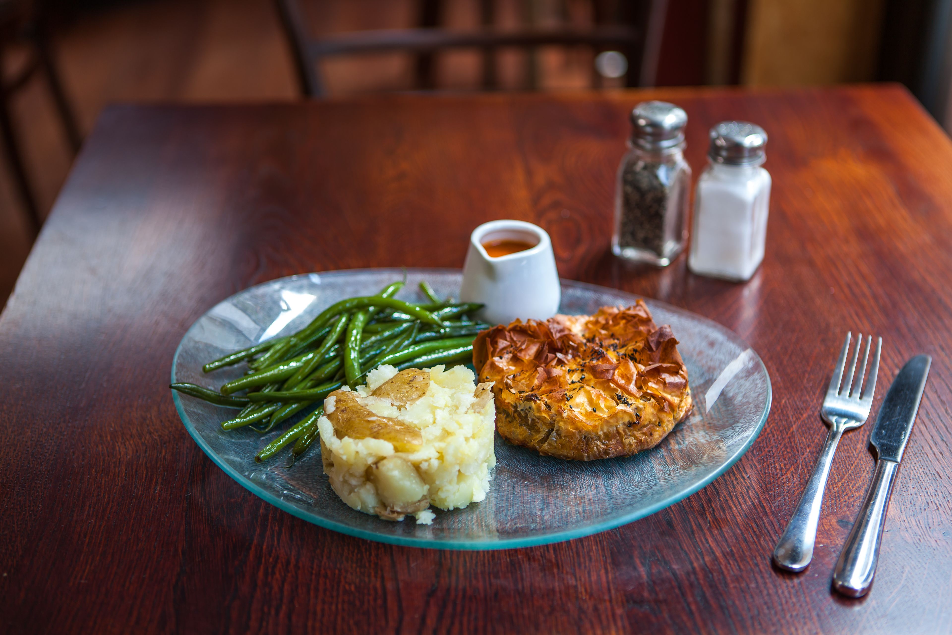 A plate with mashed potatoes, green beans, and a meat dish, accompanied by a small bowl of sauce and salt and pepper shakers on a wooden table.
