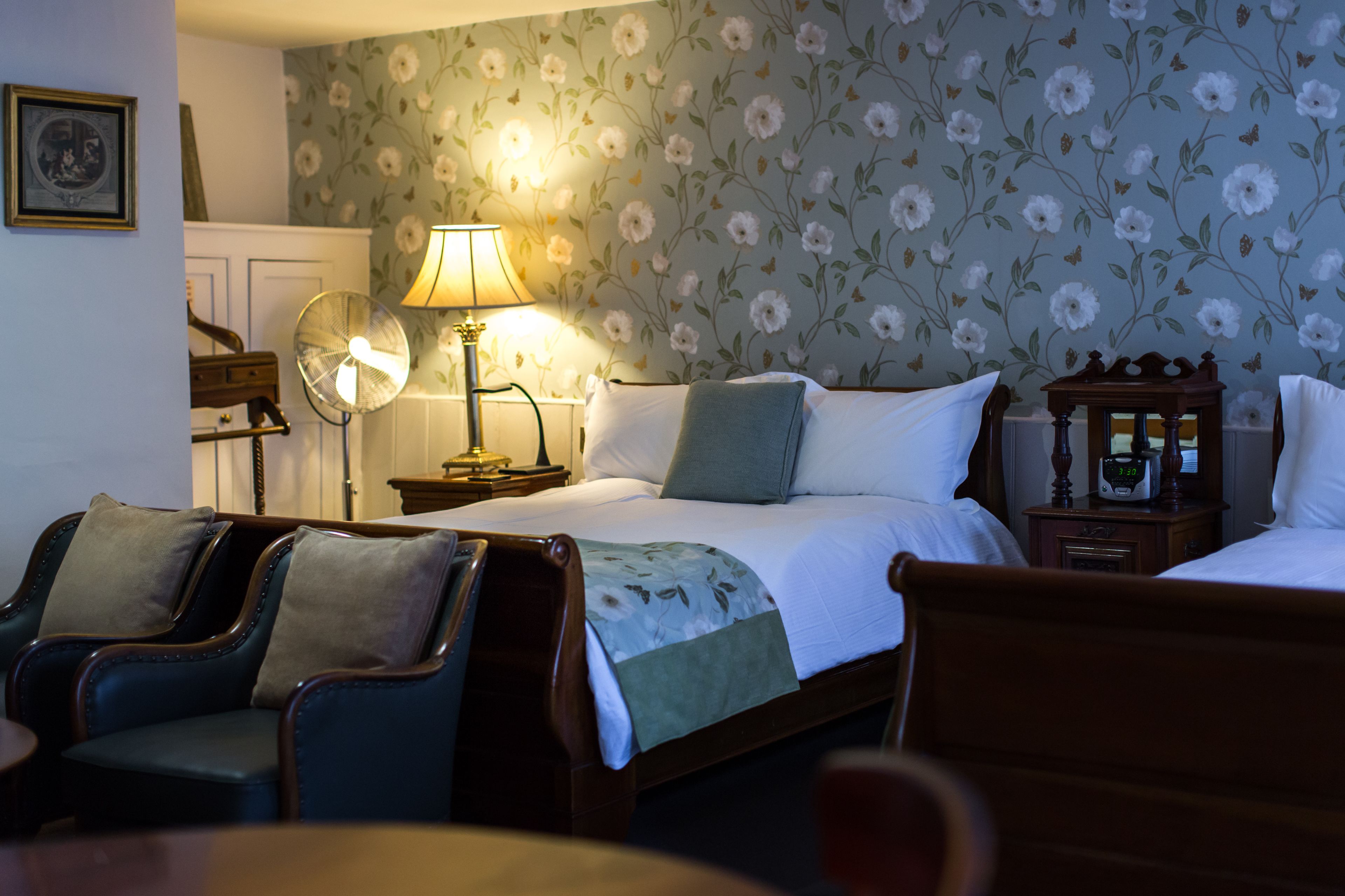 A bedroom with a bed, two bedside tables, a lamp, a fan, and two armchairs.