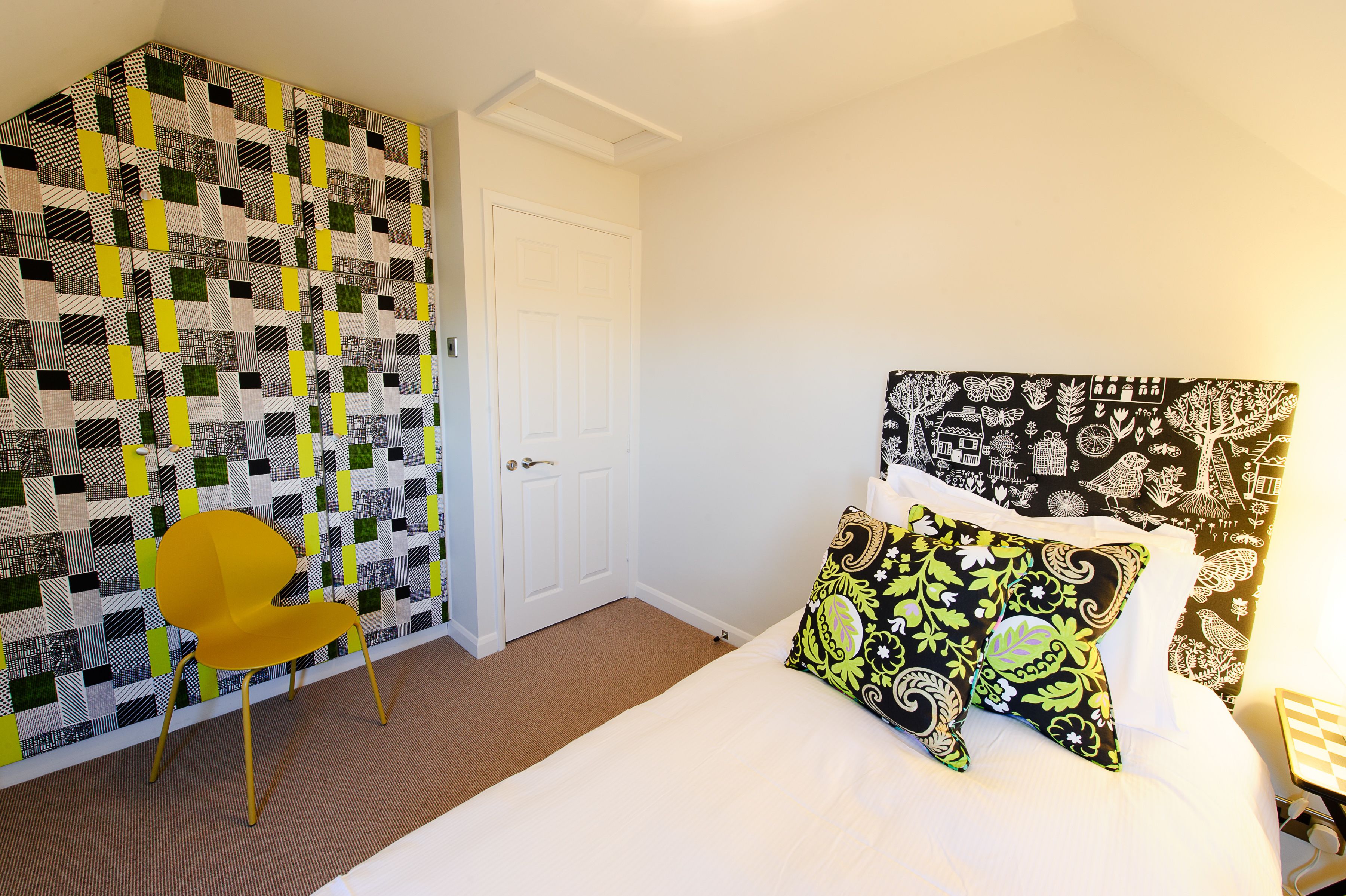 A room with a checkered wall, a bed with patterned pillows, a yellow chair, and a white door.