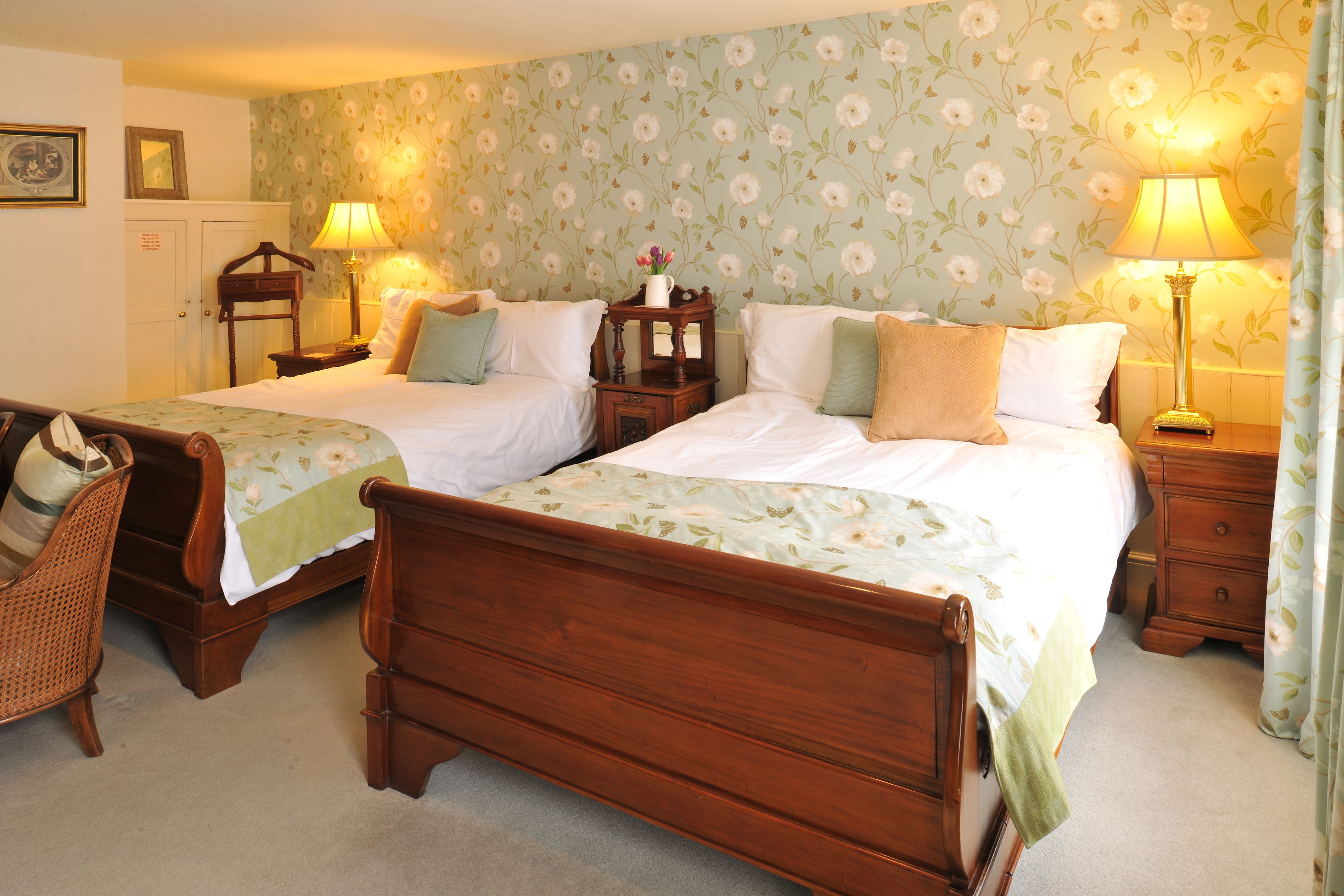 A hotel room with two wooden beds, a wooden chair, two nightstands with lamps, and floral wallpaper.