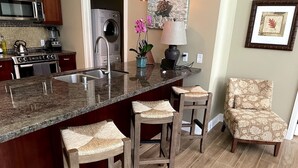 A kitchen with granite countertops, wooden stools, a microwave, a washing machine, and a decorative lamp. - Maui Resort Rentals: Groundfloor Interior Honua Kai Konea 105, Large Lawn Area, Steps to Pools/Beach/B.B.Q.s! (Lahaina)