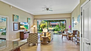 A spacious living area with a large sofa, a coffee table, and a dining table set for four. There is a ceiling fan, a kitchen area with a countertop, and a view of a lush garden through a glass door. - Maui Resort Rentals: Groundfloor Interior Honua Kai Konea 105, Large Lawn Area, Steps to Pools/Beach/B.B.Q.s! (Lahaina)