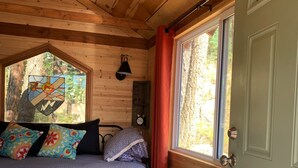 Ein gemütliches Schlafzimmer in einer Holzhütte mit einem Bett, bunten Kissen, einer Holzbalkendecke, einem Fenster mit Vorhängen und einem kleinen Tisch.