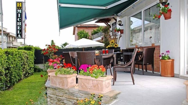 An outdoor patio with wicker furniture, a stone planter with flowers, and a green awning. - Hotel Michela (Massa)