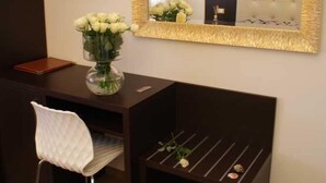 A hotel room with a dark wooden desk, a white cushioned chair, a small side table with a vase of white flowers, a large mirror with a gold frame, and a flat-screen TV mounted on the wall. - Hotel Michela (Massa)