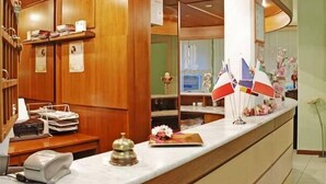 A reception area with a wooden reception desk, a telephone, a cash register, and a bell. There are flags and a flower arrangement on the counter. - Hotel Michela (Massa)